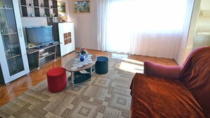 A living room with a TV, a red ottoman, a blue chair, and a large window with curtains. - Apartment Josip by Interhome (Lovran/Tuliševica)
