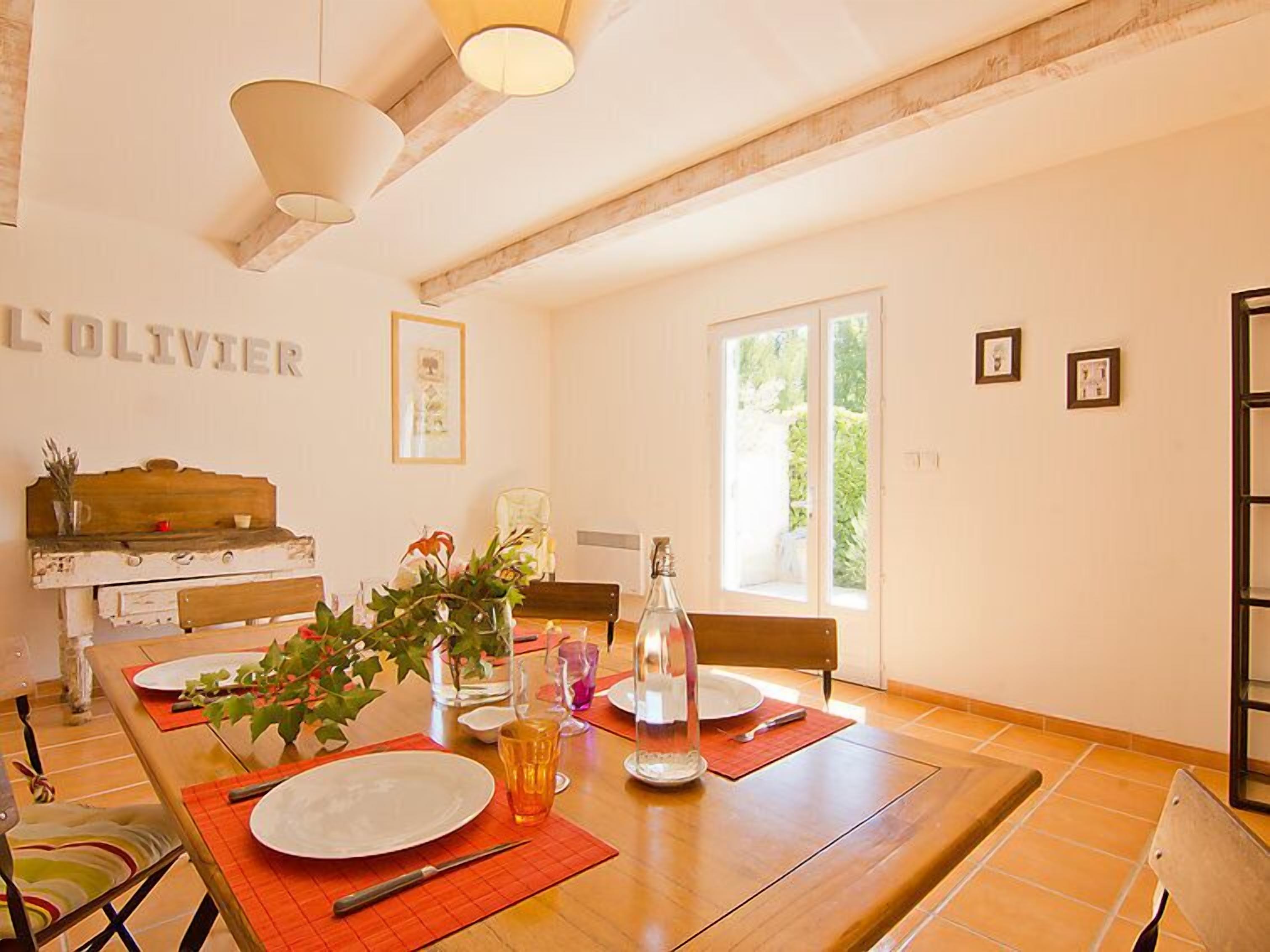 Comedor con mesa de madera dispuesta para cuatro, un florero con flores y una botella de agua.