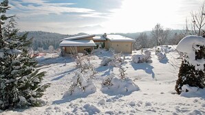 Schneebedeckte Landschaft mit einem Haus und einem Nebengebäude, umgeben von Bäumen und einem klaren Himmel.