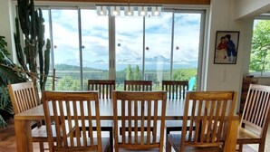 Ein Essbereich mit Tisch und Stühlen, große Fenster mit Blick auf Grünflächen und Berge sowie ein Bild an der Wand.
