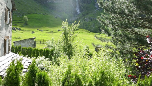 En traditionel bygning i sten med et skråt tag, omgivet af frodig vegetation og et vandfald i baggrunden.