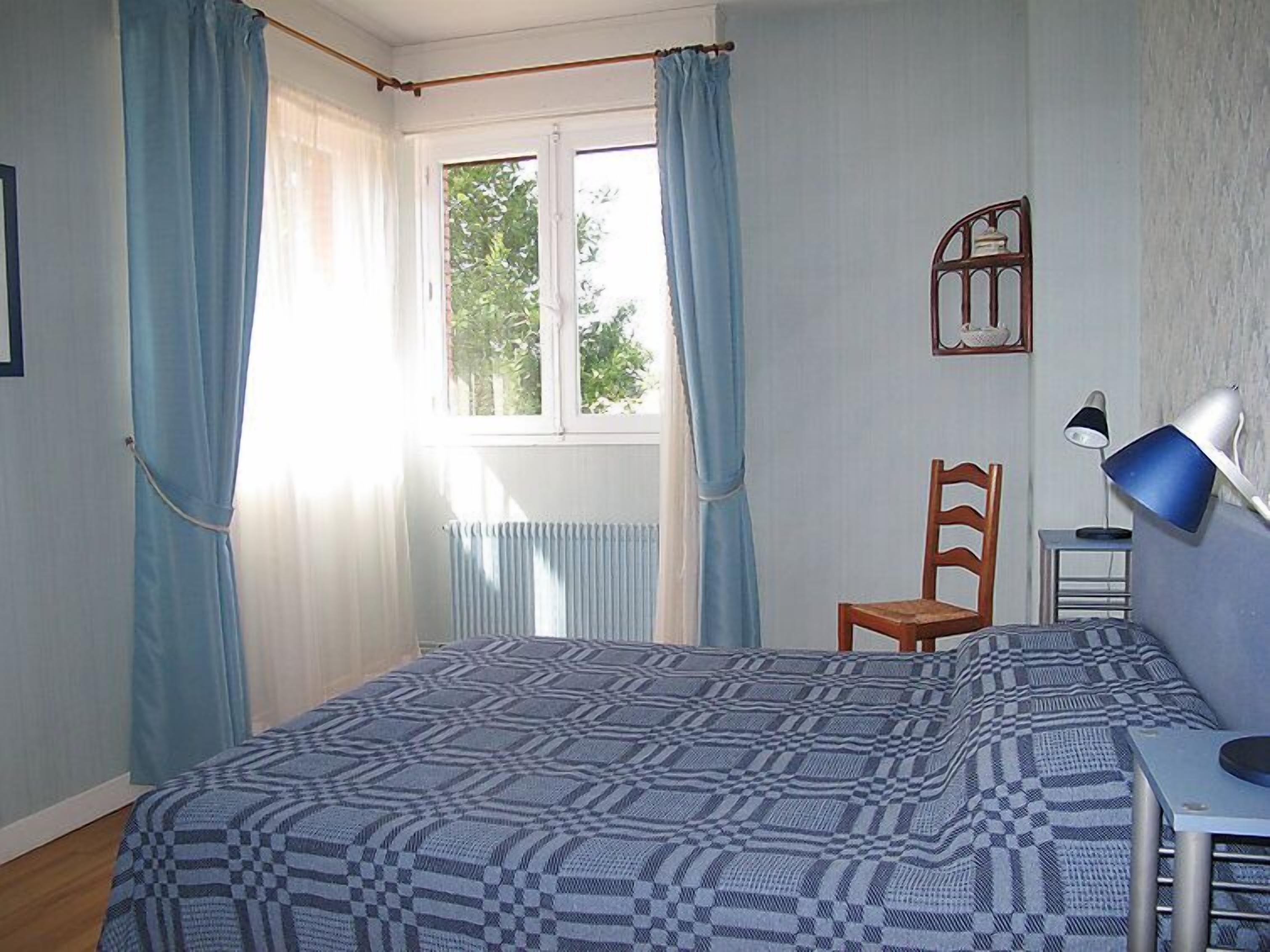 A bedroom with a bed, a chair, a desk with a lamp, and a window with curtains.