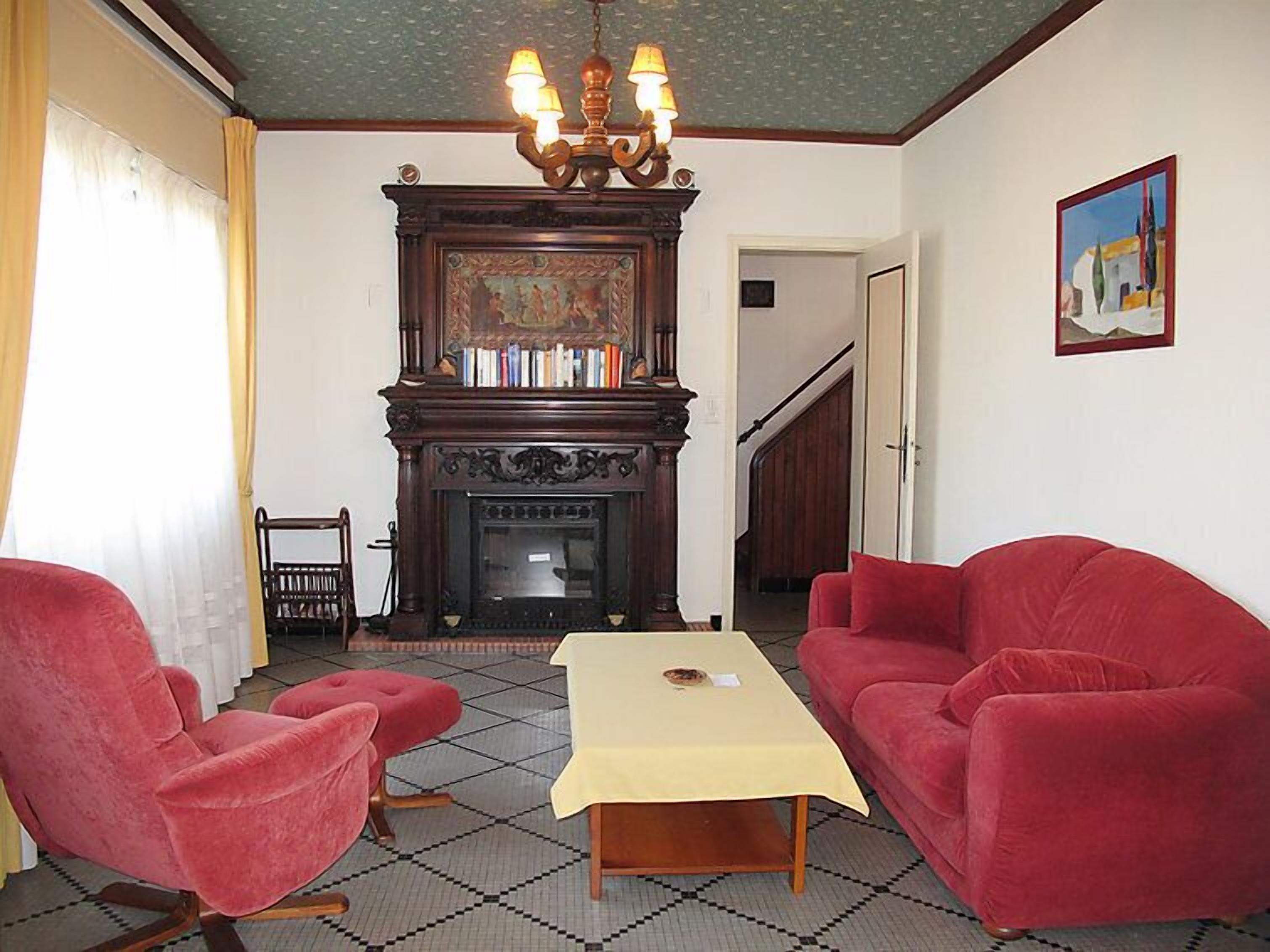 Una habitación con chimenea, dos sillones rojos, una mesa y una araña.