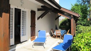 Un espacio al aire libre cubierto con dos sillones azules, una mesa de madera y una puerta que da a un espacio interior sombreado.