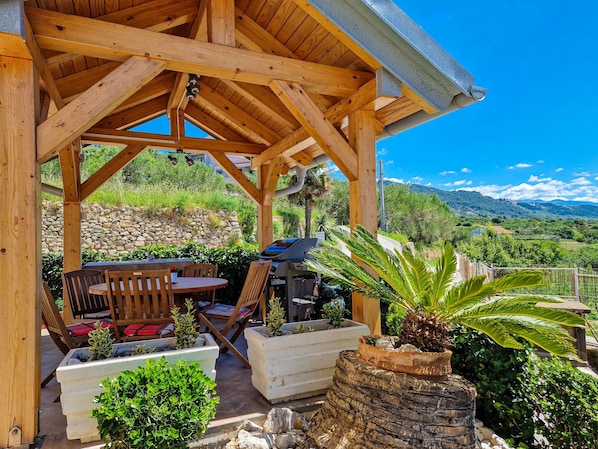 A wooden gazebo with a table and chairs, surrounded by potted plants and a stone wall.
