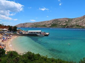 A coastal beach scene with clear turquoise waters, a docked boat, and a beach with umbrellas and people. - Apartment Olga by Interhome (Rab/Rab)