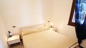 A single bed with a beige bedspread in a room with white walls, a nightstand with a lamp, and a window with a blue curtain.
