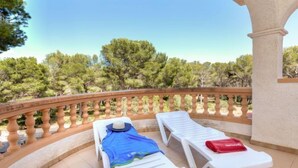 A balcony with two white sun loungers, one with a blue towel and a hat, and a red towel on the other. A woven bag is on the floor.