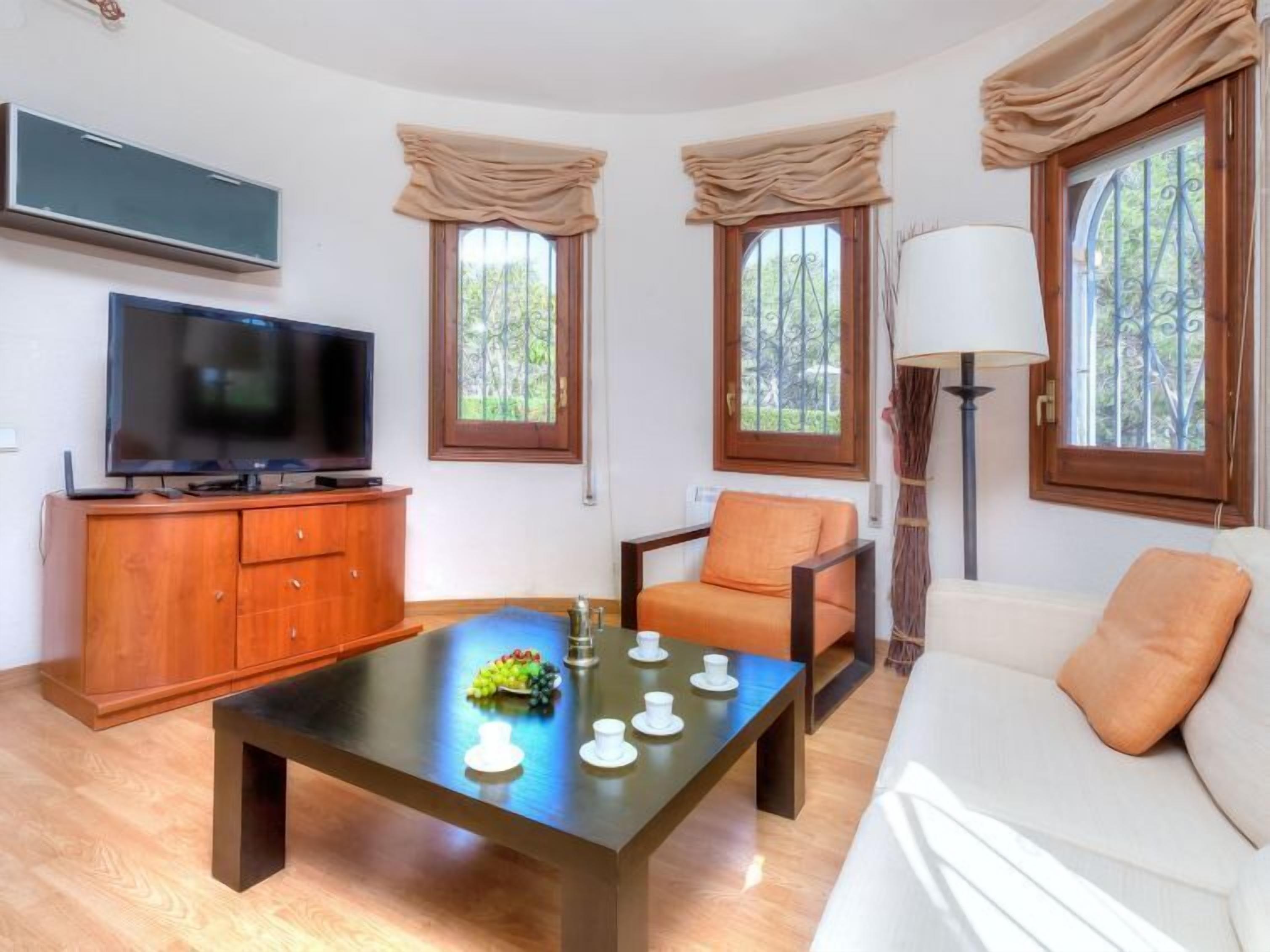 A living room with a flat-screen TV, wooden cabinet, sofa, armchair, and a coffee table with cups and a fruit bowl.