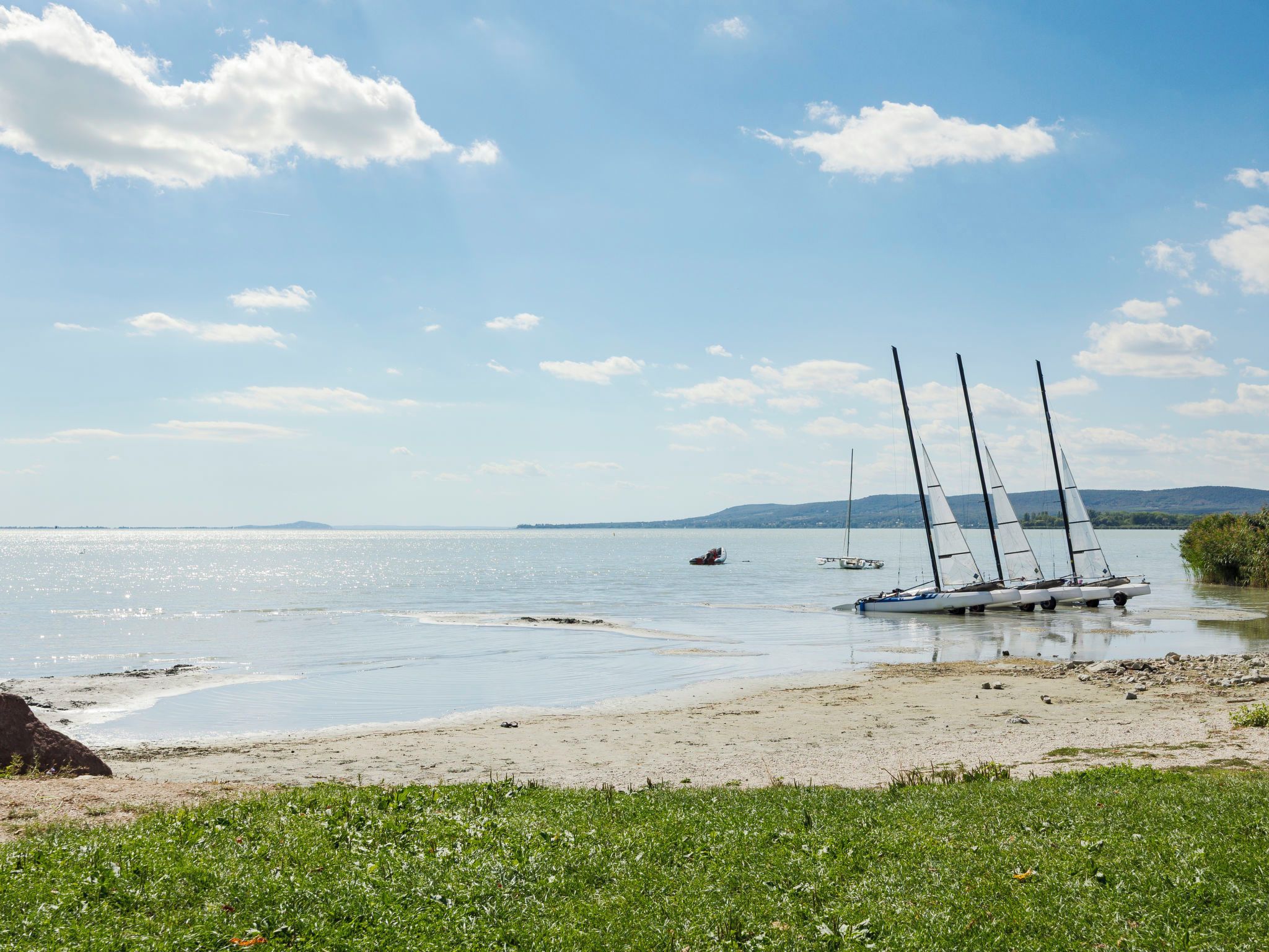 A calm lake with sailboats anchored near the shore, surrounded by greenery and distant hills.