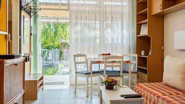 A cozy living room with a sofa, a dining table with chairs, and a TV. There is a window with curtains, a bookshelf, and a door leading outside. - Piccola by Interhome (Balatonakali)