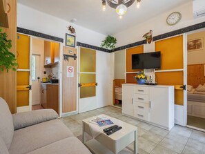 A compact living space with a sofa, a small table with remote controls, a kitchenette, a television, and a bed.