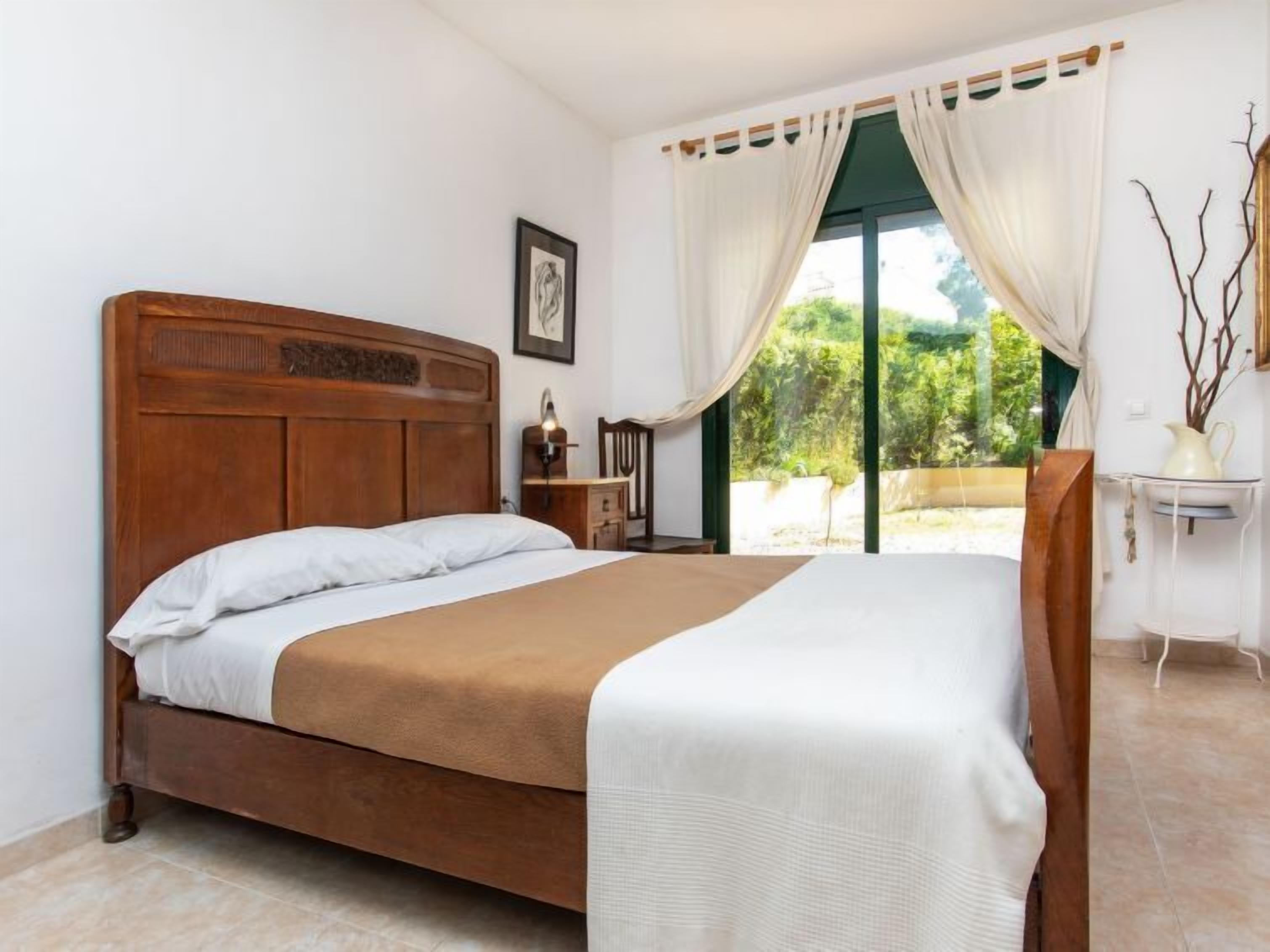 A bedroom with a wooden bed, a nightstand, a bedside table, and a view of greenery through a window with curtains.
