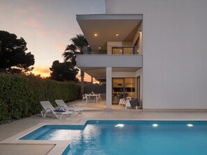 A modern two-story house with a swimming pool, outdoor seating, and a view of the sunset.