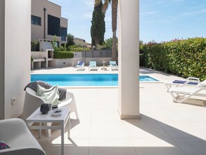 Un área de piscina moderna al aire libre con sillones de descanso y una mesa, rodeada por un seto bajo y una palmera alta.