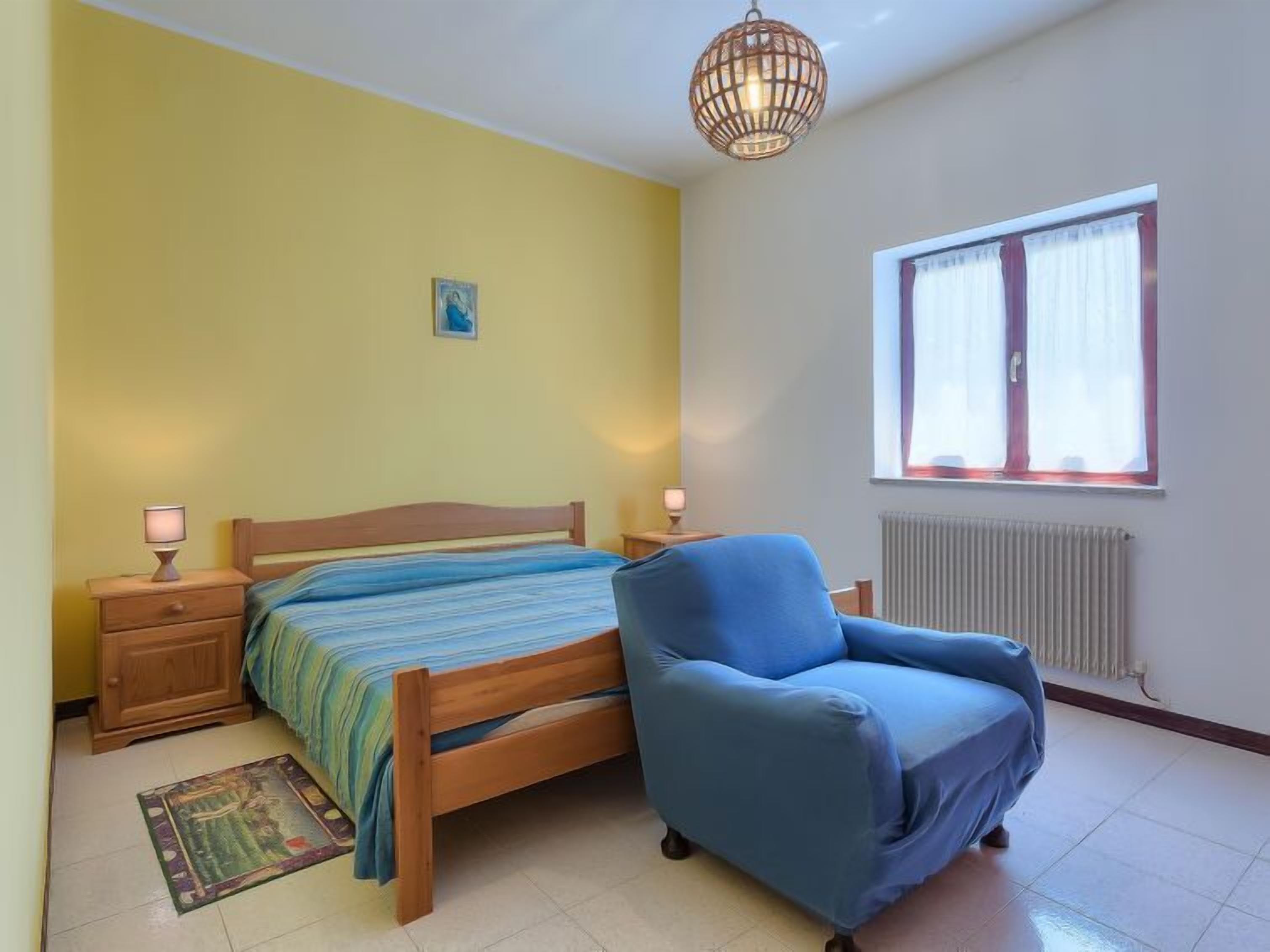 A bedroom with a bed, a blue armchair, a nightstand with lamps, a radiator, and a window with curtains.