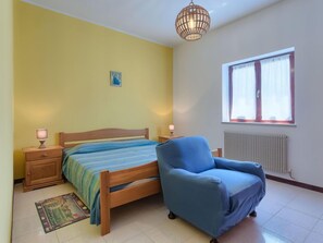 Une chambre à coucher avec un lit, un fauteuil bleu, une table de chevet avec des lampes, un radiateur et une fenêtre avec des rideaux.