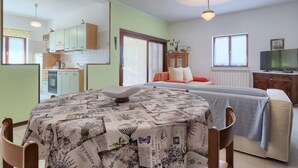 Une pièce comprenant un lit, une table recouverte d’une nappe ornée d’un motif de journaux, un téléviseur et un coin cuisine visible à travers une porte ouverte.