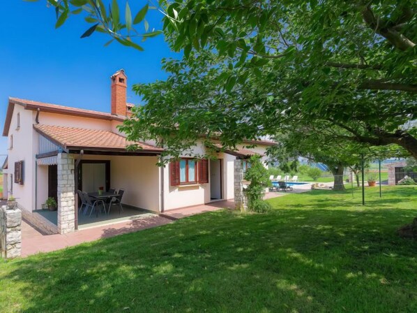Une maison à deux étages, avec un toit en tuiles, une cheminée et un patio couvert aménagé avec du mobilier d’extérieur.
