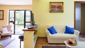 A living room with a white sofa, a television on a stand, a framed picture on the wall, and a view through a glass door.
