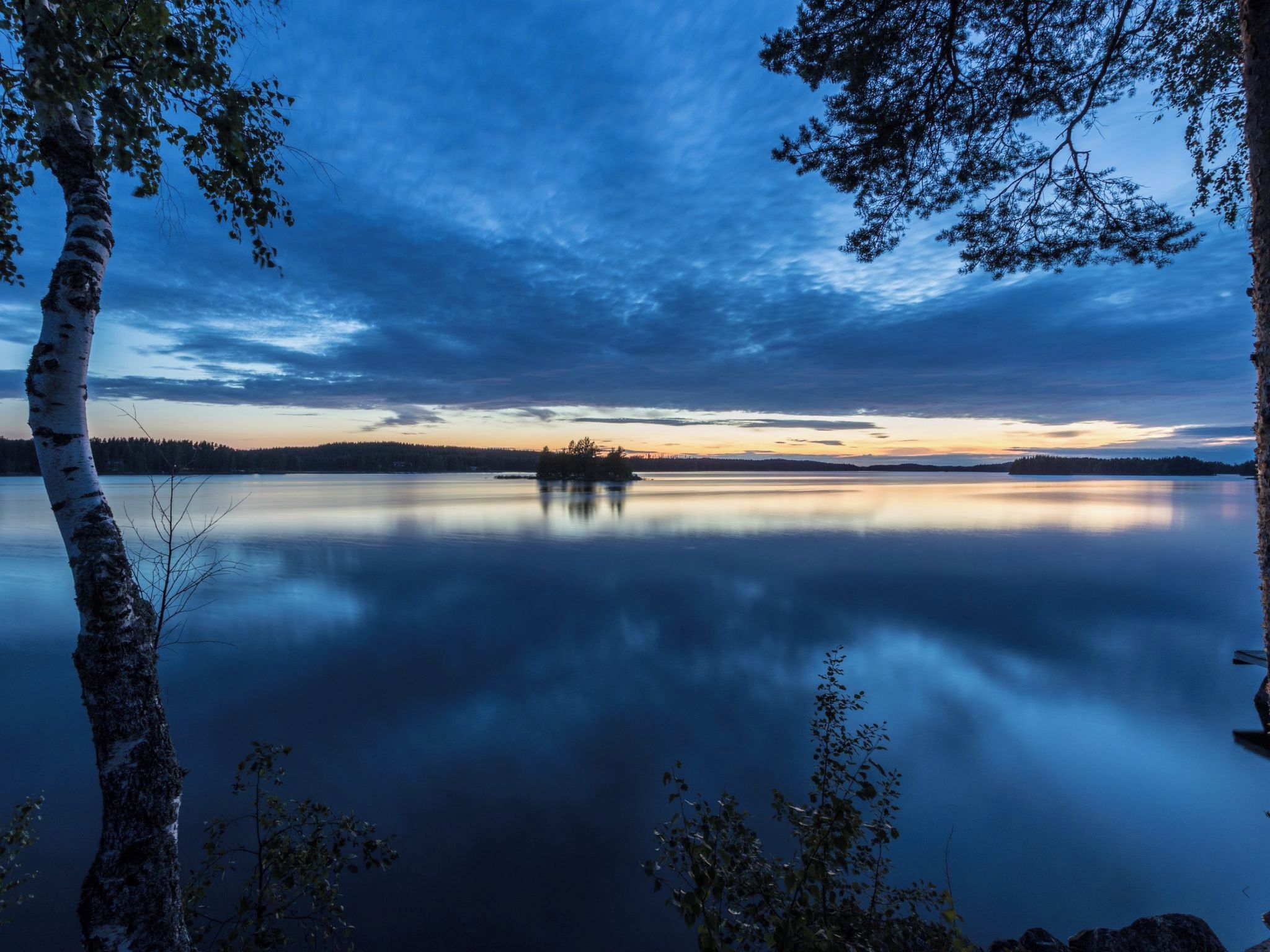 A calm lake with a clear reflection of trees and a distant shoreline.