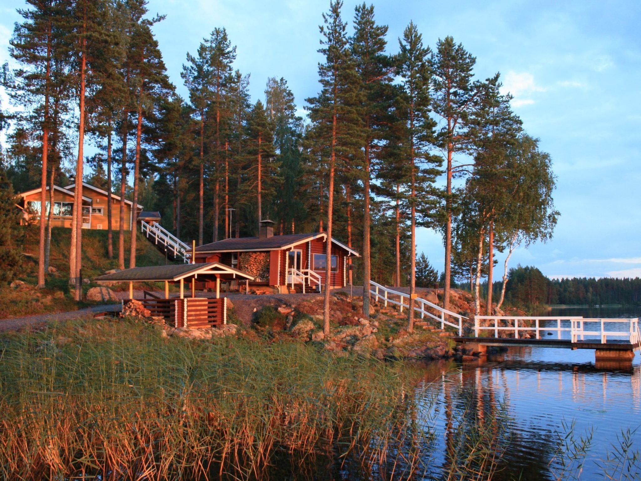 Eine Holzhütte mit einer Holzterrasse und einer Brücke über einem See, umgeben von hohen Bäumen.