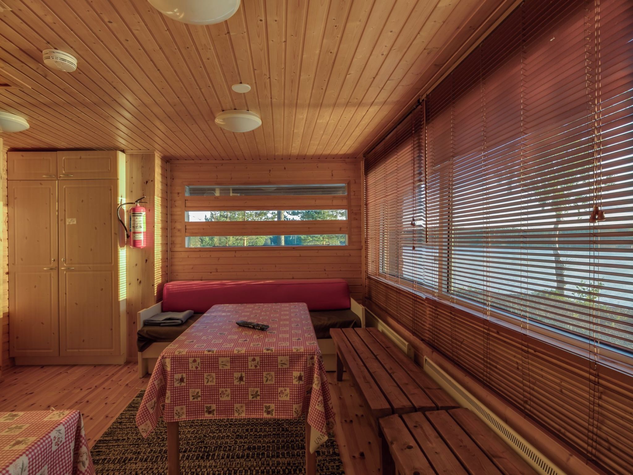 Interior de una cabaña con techo de madera, una mesa con mantel a cuadros, un banco y una ventana con persianas.