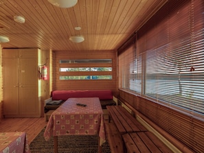 Interior de una cabaña con techo de madera, una mesa con mantel a cuadros, un banco y una ventana con persianas.