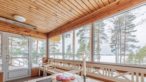 Une cabane en bois dotée d’une grande fenêtre donnant sur un paysage enneigé. À l’intérieur, on trouve une table avec une bougie, un présentoir en verre contenant des pâtisseries et un banc.