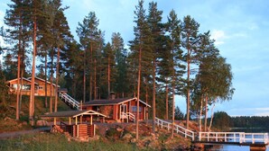 Une cabane dotée d’une terrasse et d’un escalier menant à un quai surplombant un lac paisible, le tout entouré de grands arbres.