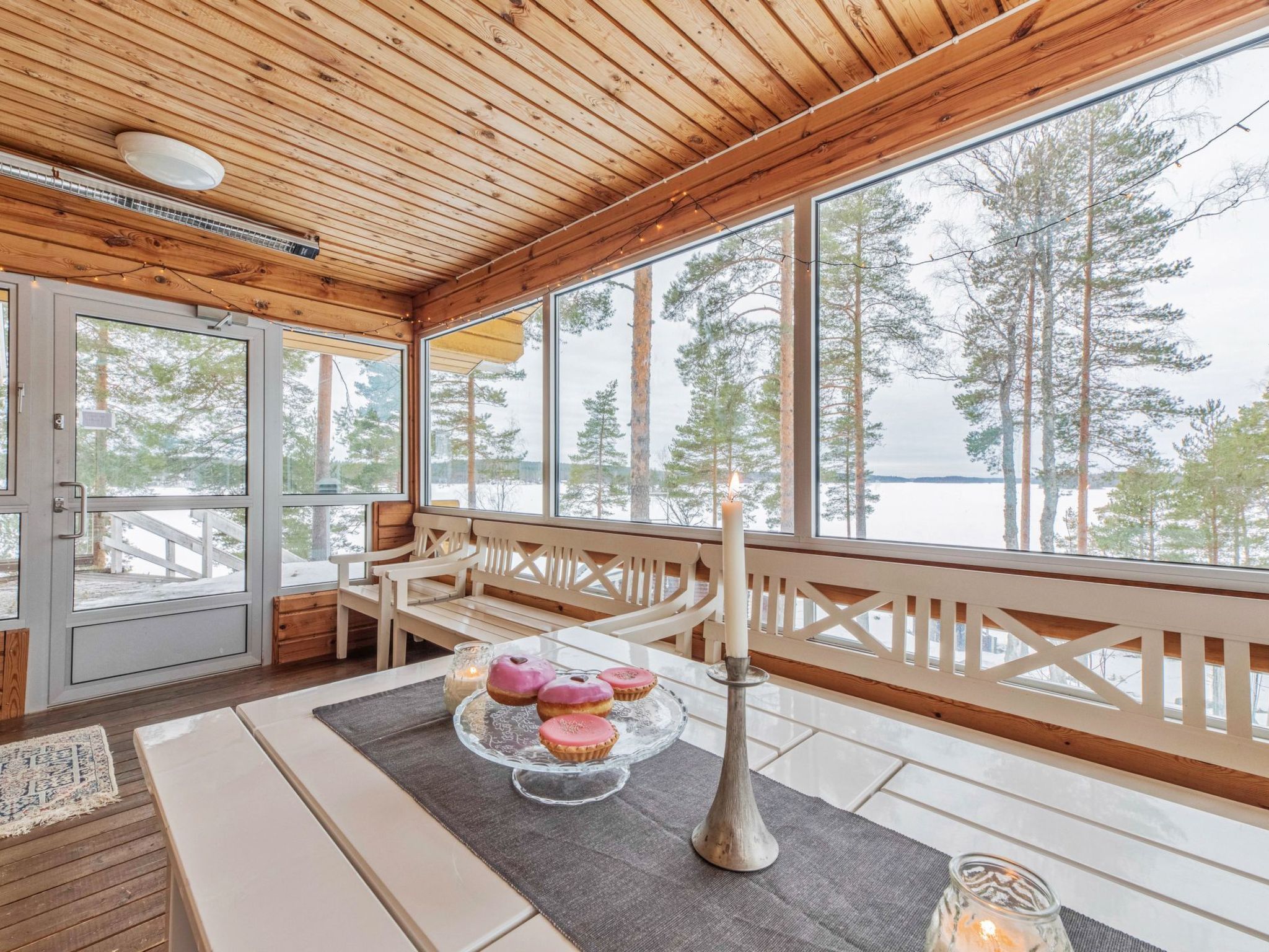 A wooden cabin with a large window overlooking a snowy landscape. Inside, there is a table with a candle, a glass cake stand with pastries, and a bench.