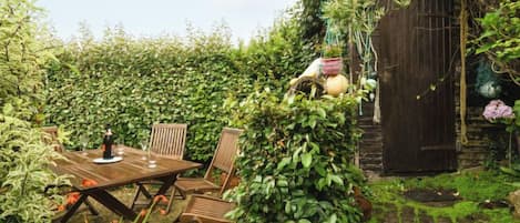 A wooden table with chairs in a garden area with dense foliage and a wooden structure in the background.