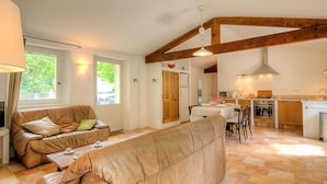 Ein gemütliches Wohnzimmer mit einer Couch und einem Essbereich, verbunden mit einer Küche mit Holzbalken und Blick ins Freie.