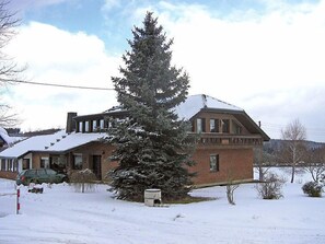 Ein zweistöckiges Backsteinhaus mit schneebedecktem Dach und einem großen immergrünen Baum davor.