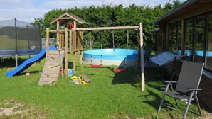 Un patio trasero con una estructura de juego de madera, un trampolín, un columpio y una piscina.