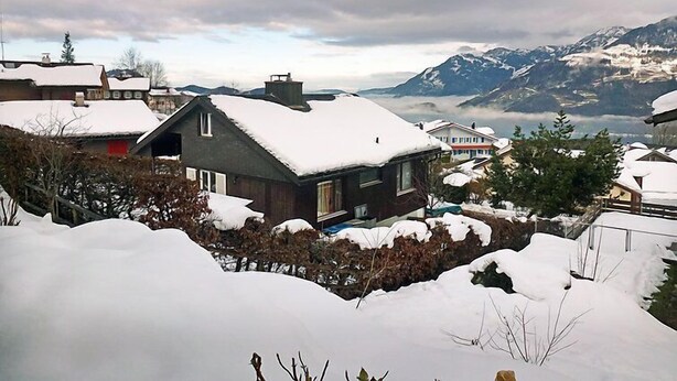 Een woonwijk met huizen onder een laag sneeuw, omgeven door bergen en een bewolkte lucht.
