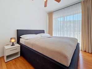 A modern bedroom with a bed, bedside table, lamp, and a view of the outdoors through sheer curtains. - Apartment Anđa by Interhome (Trogir/Slatine)