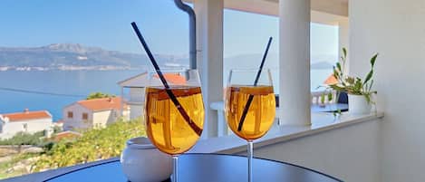Two glasses of amber liquid with straws on a round table, overlooking a coastal landscape with buildings and mountains.