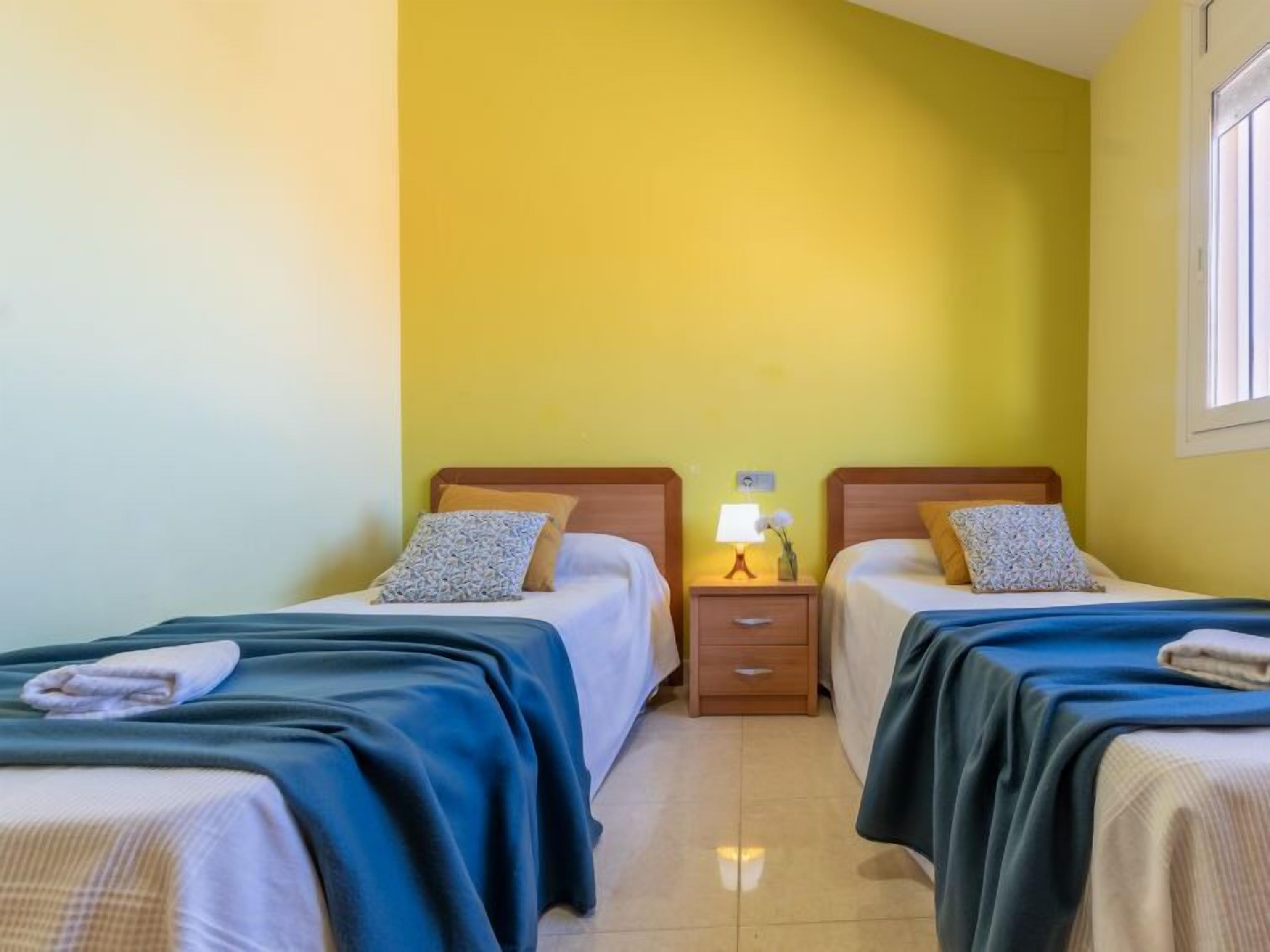 A room with two beds, each with a blue bedspread and a pillow. There is a nightstand with a lamp and a small vase with flowers between the beds.