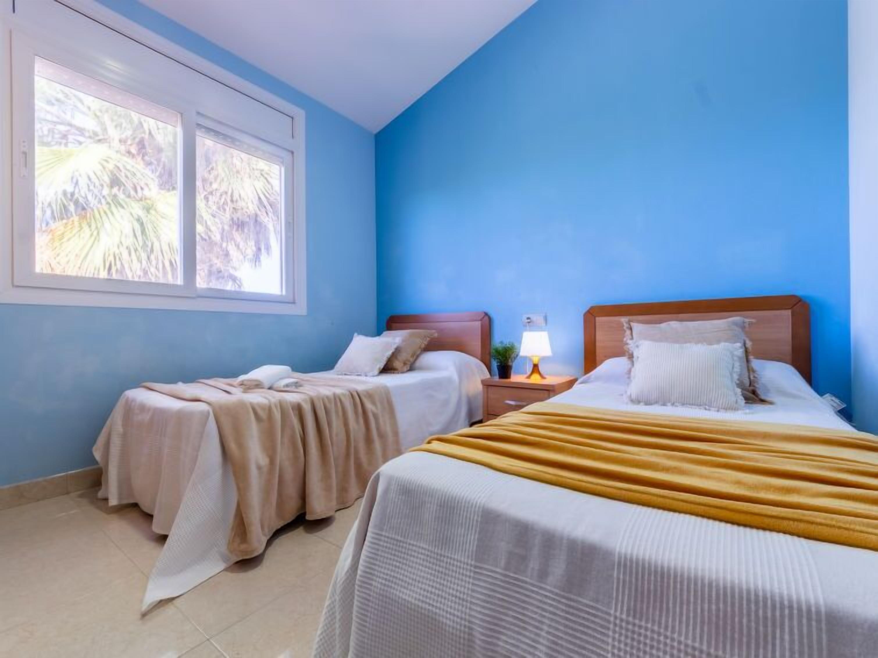 A bedroom with two beds, a window with a view of palm trees, and a nightstand with a lamp.