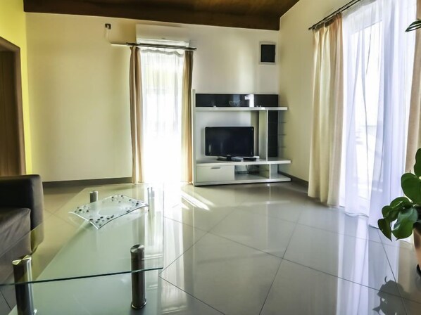 A modern living room with a glass coffee table, a flat-screen TV on a white entertainment unit, and large windows with sheer curtains.