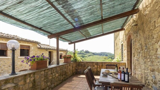 Un patio techado al aire libre con muros de piedra, una mesa de madera con sillas y plantas en macetas.
