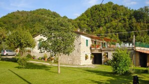 Un bâtiment en pierre doté d’un balcon, entouré d’une végétation luxuriante et avec une colline en arrière-plan.