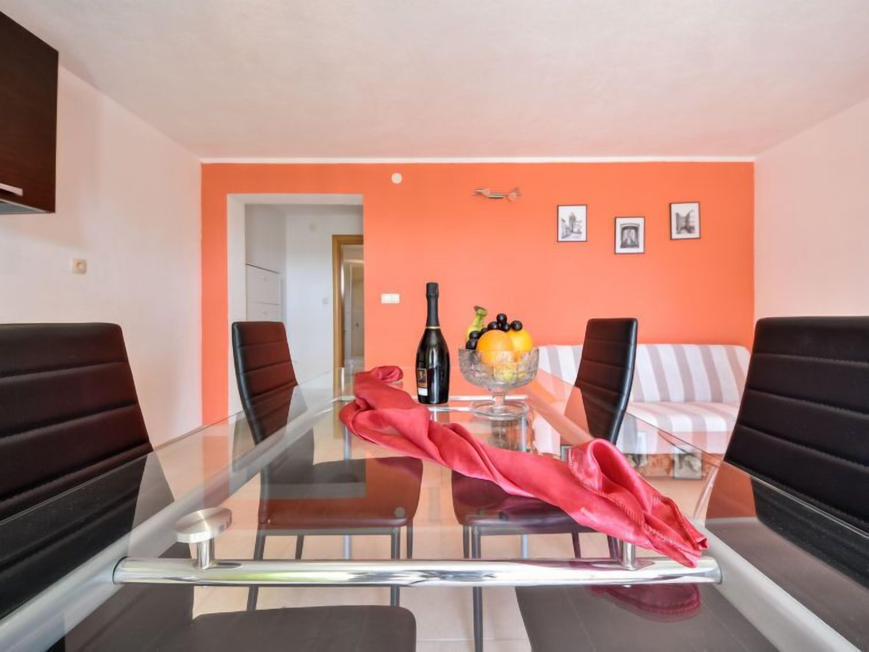 A dining area with a glass table, black chairs, a red cloth, a bottle of wine, and a bowl of fruit.