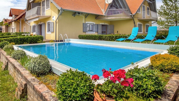 Un área de piscina con una pileta rectangular, sillas de descanso y un edificio con techo de tejas rojas y balcones.