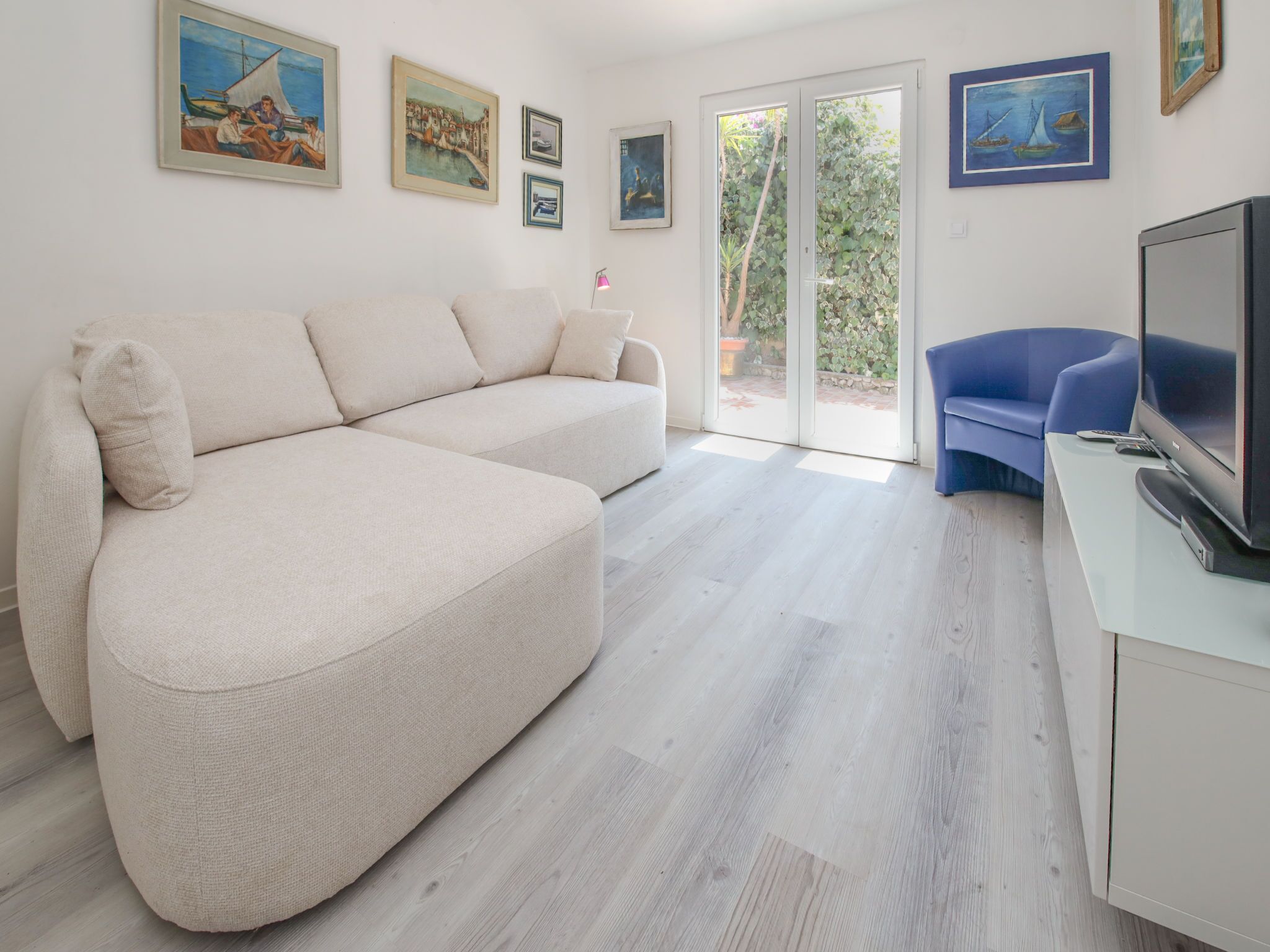 Ein modernes Wohnzimmer mit einem beigefarbenen Ecksofa, einem blauen Sessel und einem Flachbildfernseher auf einem weißen Schrank. An den Wänden befinden sich mehrere gerahmte Kunstwerke, und eine Glastür öffnet sich zu einem Außenbereich.