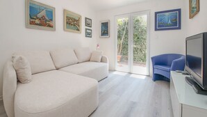 Un salon moderne avec un canapé d’angle beige, un fauteuil bleu et un téléviseur à écran plat posé sur un meuble blanc. Plusieurs œuvres d’art encadrées ornent les murs, et une porte-fenêtre coulissante donne accès à un espace extérieur.