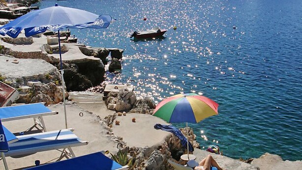 Une plage avec des chaises longues et des parasols, un terrain rocheux et un bateau sur l’eau.