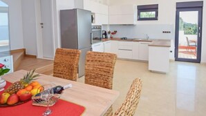 A modern kitchen with white cabinets, a stainless steel refrigerator, and a dining area with wicker chairs and a table set with fruits and a wine glass. - Vacation home Andrea by Interhome (Trogir/Okrug Gornji)