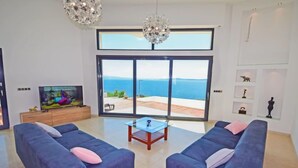 A modern living room with two blue sofas, a glass coffee table, and a large sliding glass door leading to a balcony with a view of the sea. - Vacation home Andrea by Interhome (Trogir/Okrug Gornji)